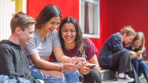 Three students sit on steps and smile at phone
