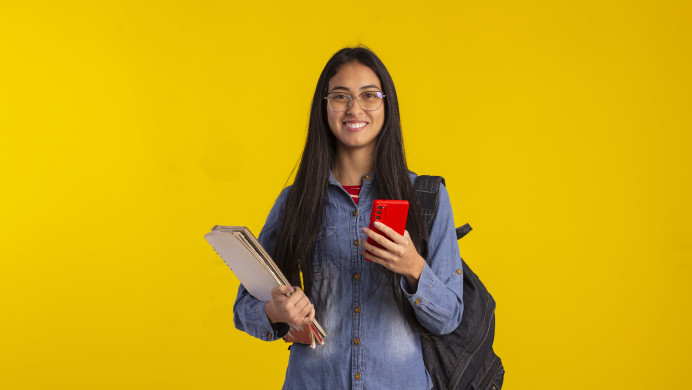A young woman stands holding a notebook and her phone in front of a bright yellow background