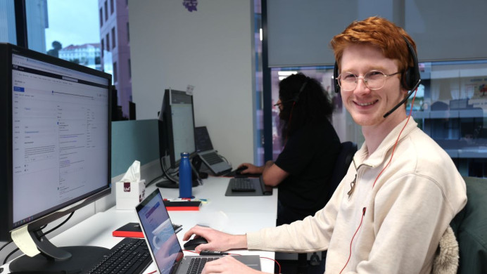 An NZQA staff members looks at the camera and smiles. He is working in an office and is sitting in front of his desk.
