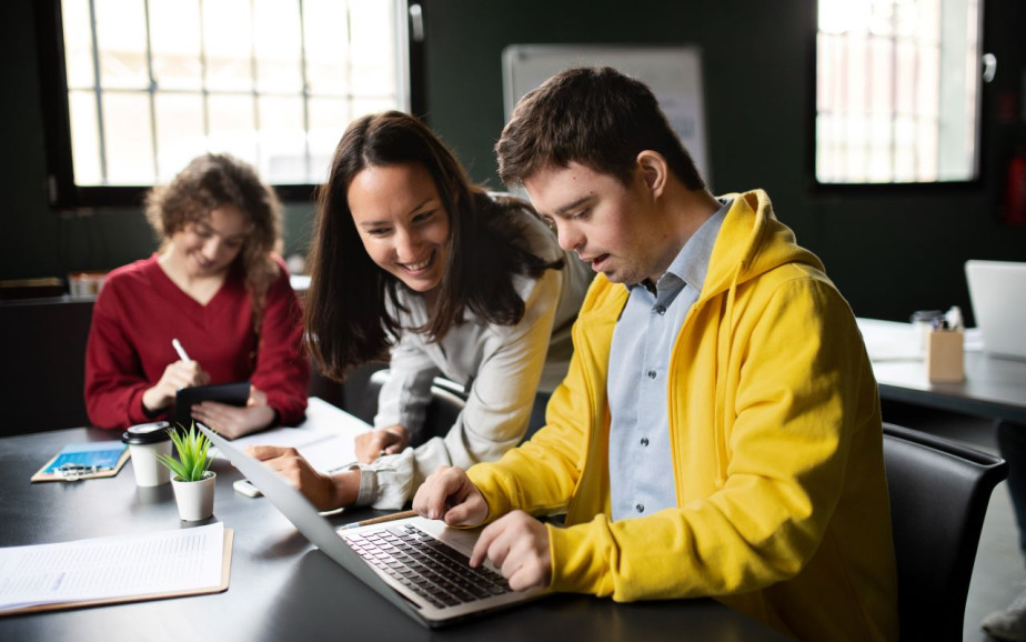 A teacher assists an NCEA student, who is working on a computer. Another student is studying in the background.