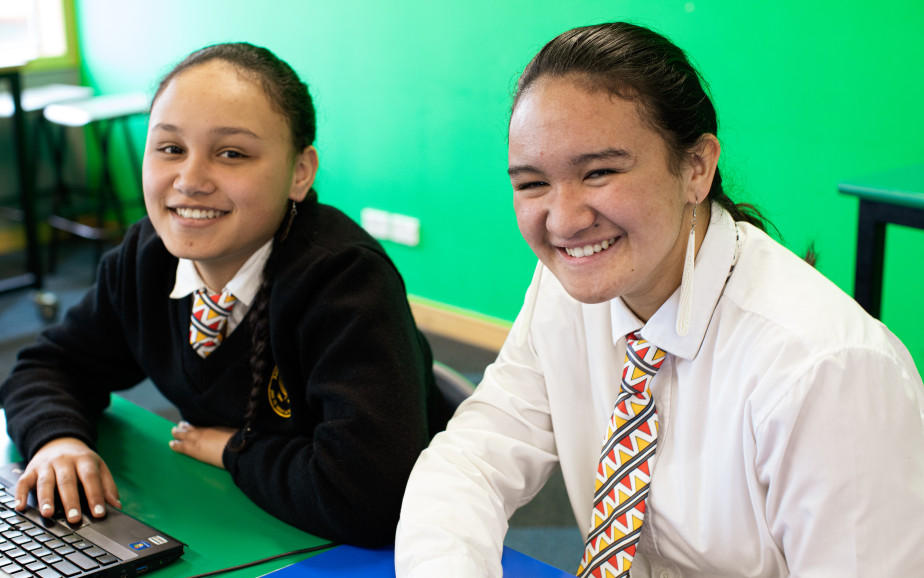 Two high school students in uniform sit in front of a green background