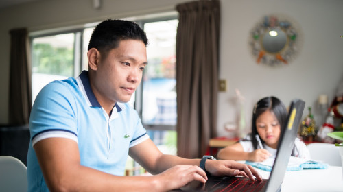 A man sits working on a laptop while his daughter sits doing school work at the same table
