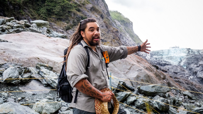 Young male Māori tour guide outside on rocky crag gestures out to sea while talking