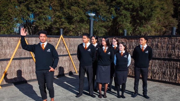 A student orates on the marae while another group of students watches.