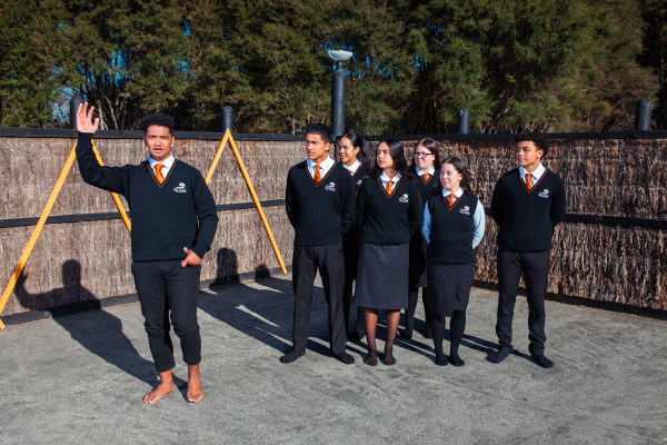 A student orates on the marae while another group of students watches.