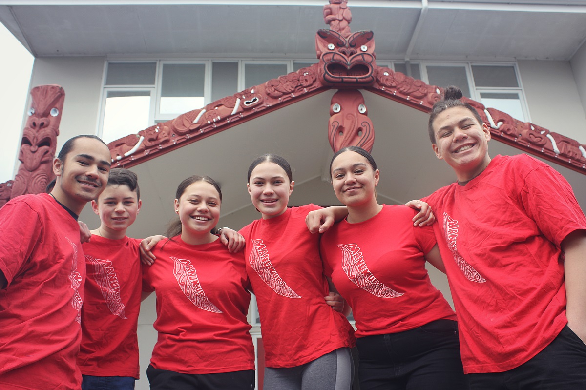 Six young people in red t-shirts smile at the camera in front of a mārae