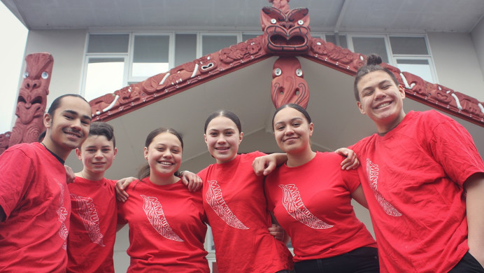 Six young people in red t-shirts smile at the camera in front of a mārae