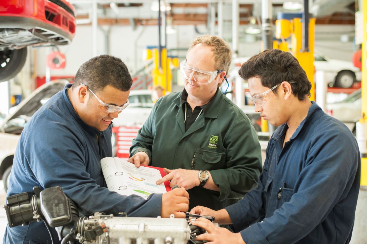 Three mechanics are looking at documentation material while working on an engine.