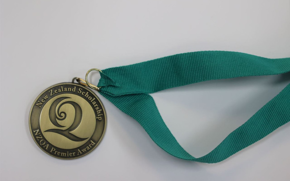 A close up image of a Top Scholar medal. The medal is gold in colour and is embossed. It has a teal ribbon attached.